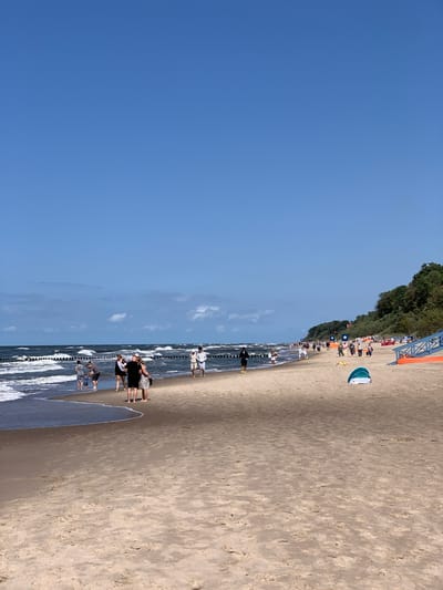 The image depicts a wide, sandy beach in Rewal, Poland, under a clear blue sky. The ocean is calm with moderate waves, and a line of wooden breakwaters stretches across the water. Several people are visible on the beach, some walking along the shore, others near the water's edge.
In the foreground, the sand is textured and appears dry, with some footprints and natural debris. Closer to the water, the sand is wet and darker. A small, light blue and white tent is set up on the beach.
Further down the beach, a group of people are gathered, and in the distance, more individuals are seen strolling. Along the edge of the beach, a densely wooded area with lush green trees forms a natural backdrop. On the right side of the image, a bright orange structure with blue railings, possibly a ramp or seating area, is partially visible. The overall atmosphere is one of a relaxed summer day at the seaside.