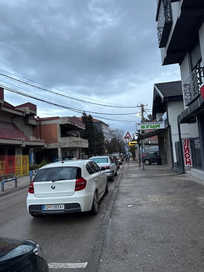 The image depicts an urban street scene in Kumanovo, North Macedonia, under an overcast sky. The ground, including the asphalt roadway and concrete sidewalk, appears wet, suggesting recent precipitation.

In the immediate foreground, the front hood and headlight of a dark-colored vehicle are partially visible on the left. Directly ahead, a white BMW 1-series hatchback, license plate KU 1183AI, is parked facing away from the viewer on the left side of the street. To its left, a line of several other vehicles, including a light-colored sedan, are parked parallel to the curb.

The sidewalk to the right of the parked cars shows damp patches and minor debris. Further down the sidewalk, an individual is visible walking away from the camera. Utility poles with multiple overhead power lines traverse the scene. A red triangular warning sign is visible on the right side of the street in the mid-distance.

Buildings line both sides of the street. On the left, several multi-story structures feature exposed concrete, red brick sections, and red sloped roofs, with a colorful yellow and red fence in front of one. On the right, a modern white and black building with ornate metal balconies is partially visible. Adjacent to it, a single-story commercial establishment displays a prominent green sign with Cyrillic text reading "БУРЕКЏИЛНИЦА БУЦИ" (Burek Shop Buci). Further down the street on the right, another building exhibits a red vertical sign with Cyrillic text "ЛИОН" (Lion).

The background features additional buildings and leafless trees under a uniformly grey, cloudy sky.