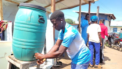 The image captures an outdoor scene in Tchere, Democratic Republic of the Congo, under bright, clear daylight. In the foreground, a young Black man, wearing a light blue and white short-sleeved t-shirt, blue jeans, and white earbuds, is washing his hands under a spigot from a large, teal-colored water barrel. The barrel sits on a rustic wooden stand. The man is focused on the task, with water visible on his hands.

Behind him, other individuals and structures are visible. To the right, another man in blue scrubs stands, while a person in a cream t-shirt and blue trousers, wearing a blue motorcycle helmet, appears to be conversing with a woman in a red top and black track pants. Two motorcycles are parked further back, one red and black, the other partially obscured.

The setting appears to be a communal or temporary area, possibly for hygiene purposes. A makeshift shelter with wooden beams and a white tarp roof extends over the foreground activity. In the background, there are various buildings and structures, including one with corrugated metal siding and a white building with blue window frames. Text partially visible on this building might read "HOPITAL GENERAL DE KAMPALA" or similar, indicating a hospital or medical facility. The man's shirt features text, likely French, including "surell arrière re" and "le aux UV", possibly indicating a brand or UV protection. A watermark "Joseph Kisuki" is present in the bottom right corner. The ground is unpaved, suggesting a natural or dirt surface.