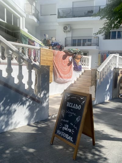 The image captures a bright, sunlit street scene in Conil de la Frontera, Spain. In the foreground, a wooden A-frame chalkboard sign displays handwritten text in white chalk advertising "HELADO" (ice cream) and its qualities: "sin azúcar" (sugar-free), "sin gluten" (gluten-free), and "NATURAL." Drawings of ice cream cones adorn the sign. Behind the sign, a white stone staircase with ornate balustrades leads up to a building. Various colorful fabrics and tapestries are displayed along the staircase and a green awning. The building itself is white with balconies and windows. The ground is paved with grey flagstones, and dappled shadows from trees fall across the scene, indicating it is daytime. There are no people visible in the frame.