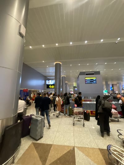 The image shows a busy airport baggage claim area. Numerous people are standing around, some with luggage, waiting for their bags. There are several baggage carousels visible, with people gathered around them. A large digital display shows "40". The overall environment appears to be a modern airport terminal with a tiled floor and a high ceiling. The specific location of Gollapalle Khurd, India, cannot be determined from this image alone.