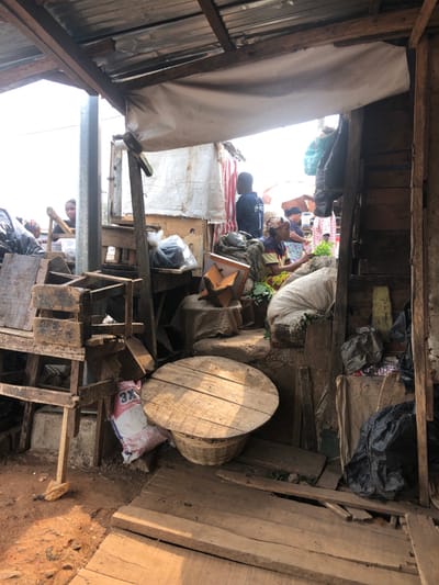 The scene depicts an outdoor market stall in a city in AlakIa, Nigeria, viewed from a partially covered interior. An overhead structure composed of corrugated metal sheeting and wooden beams is visible, with a white fabric tarp suspended beneath. The ground transitions from exposed dirt on the left to rough wooden planks on the right.
In the mid-ground, a woman is seated on the ground, wearing a striped top and patterned skirt, actively handling green leafy produce positioned near large, light-colored sacks. To her left, a wooden object covered with burlap rests on a raised surface. Behind this, a clear plastic bag containing dark material is visible on a wooden surface.
In the foreground, a stack of worn wooden crates or stools is positioned on the left. A white sack labeled "3X" in red text lies on the dirt floor. A circular wooden table top rests on a woven basket in the lower-middle portion of the image.
In the background, a person of indeterminate gender is seated behind a stall on the far left. To the right, a man wearing a dark blue shirt with the number "1" visible stands facing right. Additional market activity, including indistinct figures and stalls covered with white tarps, is visible under bright sunlight. The overall environment suggests an informal market setting constructed from wood and miscellaneous materials, with debris and black plastic bags present on the right.