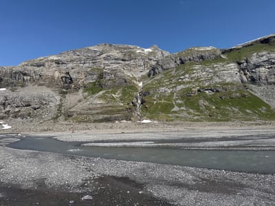 This image depicts a wide, braided riverbed filled with grey and black pebbles, with a narrow, silty grey river flowing through it. In the middle distance, a person in an orange shirt and black pants is standing near the river. Behind the riverbed, a vast, rocky mountain face rises, characterized by sharp, layered rock formations. Patches of green grass and scattered snow are visible on the mountain slopes. A thin waterfall cascades down the central part of the mountain. The sky is a clear, vibrant blue. The scene is located near the Daubensee, a lake above the Gemmi Pass, in the municipality of Leukerbad, Switzerland.
