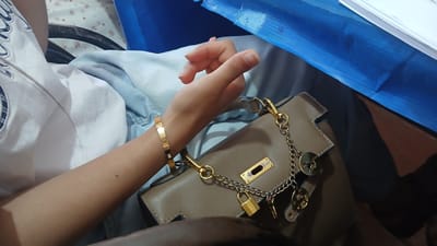 The image shows a close-up of a person's arm and hand resting on a taupe-colored handbag. The person is wearing a gold bracelet with small diamonds. The handbag has several gold charms attached to its strap, including a small padlock and circular pendants with intricate designs. The background is out of focus, but appears to be a domestic setting with a blue surface in the upper right corner. The image does not provide any information about the location being Kattakurgan City, Uzbekistan.
