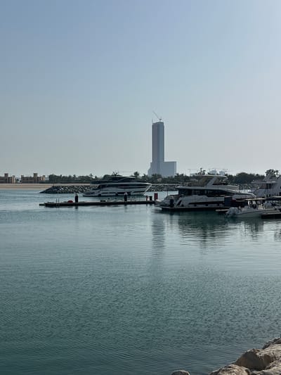 A daytime, wide shot captures a marina with several yachts docked at wooden piers. In the background, a tall, modern building under construction, topped with a crane, stands out against a clear sky. The marina is situated in the city of Al Jazirah Al Hamra, United Arab Emirates. The water is calm with subtle ripples, reflecting the sky and boats. The shoreline is lined with rocky barriers and palm trees. In the far left background, a low-rise building complex with arched windows can be seen. The scene is peaceful and portrays a developing urban coastal area.