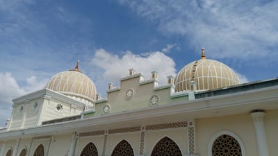 The image shows the exterior of a mosque in Julikeude Dua, Indonesia. The mosque features a large, light-colored dome with white latticework and a golden finial. Another smaller dome is visible on the left side of the building. The mosque's facade is adorned with intricate geometric patterns and decorative elements, including arched windows with elaborate latticework. The building has a light-colored exterior with white trim and green accents on the upper portions. The sky above is blue with scattered white clouds. The image is taken from a low angle, emphasizing the grandeur of the mosque. No people are visible in the image.