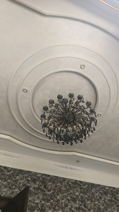 The image shows a chandelier hanging from a decorative ceiling in what appears to be an interior space. The ceiling features concentric circles and molding, with small recessed lights. The chandelier itself is dark and elaborate, with multiple tiers of crystals. Below the ceiling, a portion of a wall with a marbled pattern is visible, along with the corner of a wooden structure. The scene is well-lit, suggesting an indoor setting. No people or specific actions are depicted. The location of Shamsiabad, Uzbekistan is not visually ascertainable from this image alone.