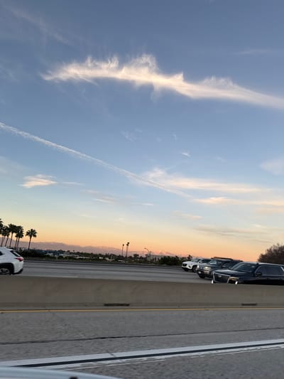 A highway view features cars parked along the roadside. In the distance, mountains rise with hints of snow. The sky displays light blue tones blending into yellow and orange hues near the horizon. Wispy clouds stretch across the sky, accompanied by an airplane contrail. Palm trees line the horizon.