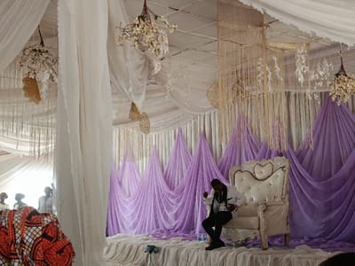 This image depicts an indoor event stage in Katugal, Nigeria, elaborately decorated for a formal gathering. The elevated stage is draped in white fabric, featuring a large, ornate white throne-like chair with a tufted back. A male individual, dressed in a white suit jacket, black vest, black trousers, and a white shirt, is seated sideways on the chair, leaning forward with his right hand near his torso, possibly adjusting his clothing or interacting with an unseen element. Two small clear bottles and a vase containing blue flowers are visible on the stage floor near his feet. The backdrop consists of vertically pleated lavender sheer fabric, forming a series of pointed peaks. The ceiling and upper portions of the walls are extensively draped with white sheer fabric, from which decorative elements are suspended. These include arrangements of white artificial flowers, golden filigree leaf-shaped ornaments, and long, cascading chandeliers made of thin, light-colored strings or beads. In the far left background, several indistinct figures, primarily women, are partially visible, suggesting attendees or participants, with a brightly patterned fabric visible in the immediate lower-left foreground.