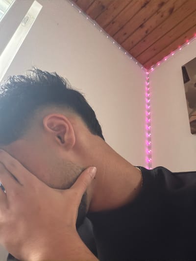 The image displays an unidentified individual, likely male, from a partial, upward-angled perspective. The individual has dark hair with a fade haircut and visible stubble along the jawline. A dark-colored shirt is worn, along with a thin gold-colored chain around the neck. The subject's left hand is positioned over the face, obscuring the eyes and upper cheek, with the thumb near the left ear. A digit, likely from the individual's right hand, is gently touching the right side of the neck/jawline area, near a small, dark mole.

The background shows an indoor environment. The walls are light-colored. The ceiling comprises a flat white section and a sloped, wood-paneled section. Pink LED string lights are affixed along the intersection of the walls and the wooden ceiling, and also vertically down a wall corner. A portion of a window frame is visible in the upper left. An indistinct, dark object, possibly a framed item, is partially visible on the right wall. The image provides no specific visual cues to identify the location as Tuttlingen, Germany.