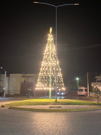 The image depicts a nighttime street scene in Las Grutas, Argentina, centered on an illuminated conical structure resembling a Christmas tree. This structure, composed of bright yellow-white lights on a metal frame and topped with a finial, stands within a circular grassy median. A tall, dark street light pole with a bifurcated top supporting two luminaires is positioned directly behind the illuminated tree. The foreground consists of asphalt pavement, reflecting the tree's lights, separated from the median by a yellow curb.

To the left of the median, a light-colored building, possibly beige or cream, with a darker reddish-brown structure attached, is visible. Further down this street, a smaller street light is illuminated. To the right of the median, a white passenger van is parked or moving slowly, its rear visible. Beyond the van, a building with a "Corona" sign is partially visible. Another vehicle with illuminated headlights is discernible further down the street on the right. The sky is dark, indicating night, with faint, distant lights visible in the far background. No individuals are explicitly identifiable in the scene.