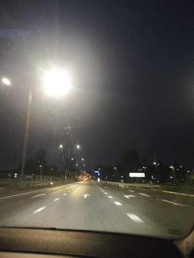 The image depicts a nighttime view from inside a vehicle driving on a multi-lane highway in Laagri alevik, Estonia. The sky is dark, with a bright streetlamp dominating the upper left quadrant, casting a significant glare. The road ahead is well-lit by numerous streetlights lining both sides. White lane markings are clearly visible, indicating multiple lanes of travel, with some showing motion blur due to the vehicle's movement. Trees and some roadside structures are dimly visible in the background. There are no people or other vehicles clearly discernible.