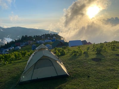 A tent is pitched on a grassy hillside overlooking a village nestled in a valley. The village, characterized by its clustered houses with diverse roof colors, extends across the rolling landscape. In the background, misty mountains rise, partially obscured by clouds. The sun, peeking through a thick layer of clouds, casts a warm, golden light across the scene, suggesting it is either early morning or late afternoon. The sky is a pale blue, with streaks of white clouds adding texture. The overall atmosphere is serene and peaceful, evoking a sense of natural beauty and tranquility in a mountainous, rural setting.