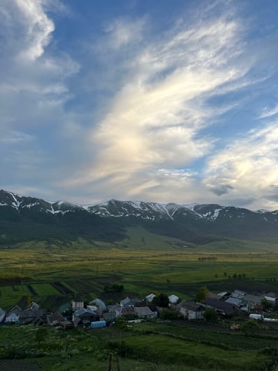 The image depicts a rural landscape in Fioletovo, Armenia, dominated by a vista of snow-capped mountains under a dramatic sky filled with wispy clouds. In the foreground, a cluster of houses forms a small village, nestled amongst green fields and agricultural plots. The houses appear to be of traditional design, with varied roofing materials and modest structures. The fields showcase distinct patterns of cultivation, suggesting ongoing agricultural activity. The mountains, likely part of the Pambak Range, rise majestically in the background, their peaks dusted with snow, contrasting with the lush green foothills and valley below. The lighting suggests either early morning or late afternoon, with the sun casting a warm glow on parts of the sky and landscape. There are no people or animals visible in the image, and the scene conveys a sense of quietude and natural beauty.