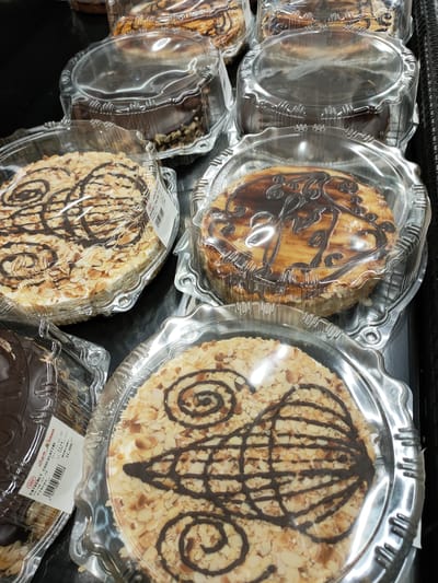 This media file shows an overhead view of several round cakes displayed in clear plastic containers. The cakes are arranged on a dark surface, likely a shelf in a store.

The cakes appear to be of different varieties. One prominently featured cake is covered in sliced almonds and decorated with a dark chocolate drizzle in a swirling pattern. Another cake, partially visible behind it, is also adorned with chocolate icing and what seem to be chocolate shavings or nuts on its sides. Further back, more cakes are visible, some with a striped chocolate topping and others covered in what looks like powdered sugar or a light frosting.

A label is partially visible on the side of one of the cakes, showing text in French and Arabic, including "ENTREMET CHOCOLATINE" and "جيان Geant". The label also includes dates, prices, and barcodes. The lighting suggests an indoor setting, possibly a bakery or a grocery store's dessert section. No people are visible in the frame. The overall impression is a selection of pre-packaged desserts.