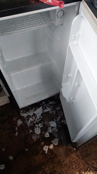 An open, empty compact refrigerator is depicted from an overhead perspective, displaying its white interior with two wire shelves and a circular thermostat control. The interior surfaces show signs of discoloration and moisture. A red plastic spoon rests on the top edge of the refrigerator, partially visible. The dark, wet, and stained floor directly beneath the unit is scattered with numerous broken pieces of white ice and frost, several exhibiting a distinct grid pattern. No individuals are visible. The specific location within Tlaxcala City, Mexico, is not discernible from the visual evidence.