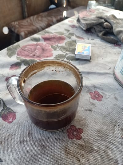 A close-up, eye-level shot shows a glass mug containing dark liquid, likely coffee, with sediment visible on the inner rim. The mug sits on a tablecloth with a faded floral pattern, predominantly pink roses. To the right of the mug, a matchbox with a colorful design is partially visible. Further to the right, a pair of work gloves and a blurry cylindrical object, possibly a container, are present. In the background, a wooden surface and a blurred chair with a patterned upholstery can be seen. The overall scene suggests a domestic or workshop setting.