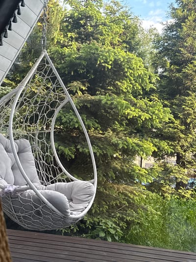The image depicts a hanging chair situated outdoors, possibly a patio or backyard, with lush green trees forming the backdrop. The chair is white with a net-like pattern and soft grey cushions. Adjacent to the chair is a dark wooden structure, likely a wall or fence. A glimpse of blue sky is visible through the trees. The location is specified as Чёрное, Russia.
