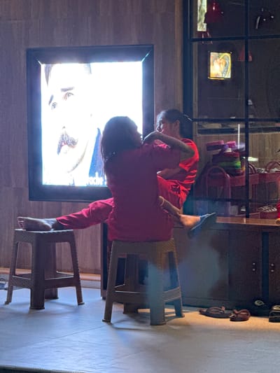 A woman and a child, both in red clothing, are perched on plastic stools indoors in Pererenan, Indonesia. The woman is helping the child adjust a large framed picture on a wall that features a close-up of a face. Behind the woman, a rack holds various items, and a picture frame is visible. A pair of shoes sits on the floor. The room is lit by natural light.