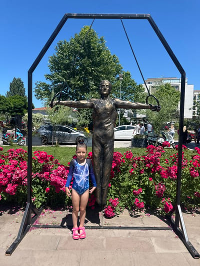 The image features a young girl posing next to a bronze statue of a gymnast holding rings. The statue is positioned in front of a vibrant bed of pink roses. The girl, wearing a blue and white leotard with a butterfly design and pink sandals, is smiling at the camera. Behind the statue and the girl, a street scene is visible, with cars and trees. In the background, modern buildings can be seen under a clear blue sky. The overall atmosphere suggests a sunny day, likely during the daytime, in a public outdoor space like a park or square. The setting, identified as Sancaktepe, Turkey, implies a specific geographical and cultural context. The image captures a moment of playful interaction between a child and a public monument, set against a backdrop of urban life and nature.