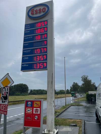 This image depicts an Esso gas station in Alsfeld, Germany, on a cloudy, rainy day. The primary subject is the tall, illuminated gas price sign displaying fuel types and their corresponding prices in red digital numbers. The prices are as follows: Truck Diesel at 1.859, Diesel at 1.869, Super E10 at 1.819, Super at 1.99, Supreme+ at 1.979, CNG at 1.359, and AdBlue at 1.359.

To the left of the gas price sign, a yellow diamond-shaped road sign is visible, along with a red sign indicating "VIDEO ÜBERWACHUNG" (Video Surveillance) and a clock icon showing "24h". Below the price sign, a red display board shows various logos, including Esso Card, DKV, and two other illegible logos.

The environment is an outdoor setting with a wet asphalt road and parking lot, indicating recent or ongoing rain. A guardrail runs along the side of the road. In the background, there are trees and a slight incline of land. Further down the road, several vehicles are visible, including a green truck, a dark car, and other vehicles in the distance. A white van with the word "HERMES" partially visible on its side is parked on the right side of the image. The sky is heavily overcast with grey clouds. There are no people visible in the frame.