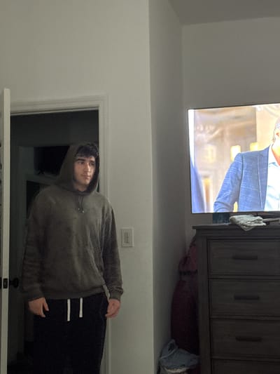 A young man stands in a doorway looking off to the side, wearing a dark green hooded sweatshirt and black sweatpants. A television is visible in the background, displaying a man in a blue checkered suit. To the right of the television is a dark dresser. The room appears to be a bedroom in a residential setting. The image was taken in Bellmore, United States of America.