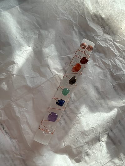 The primary subject is a clear, rectangular mineral stick, resembling selenite, adorned with seven small, tumbled, colorful stones arranged in a linear sequence. These stones, from left to right, appear to be reddish-brown, orange, dark brown/black, green, blue, purple, and clear. They are secured to the mineral stick by intricate copper wire wrapping, which includes decorative spirals at the left end of the stick.
There is no specific activity or event taking place; the image is a still-life photograph of the crafted object.
The setting consists of a crinkled, light-colored paper background, likely white or off-white tissue paper, which displays various folds and textures. Bright, natural lighting, possibly sunlight, illuminates the scene, casting distinct highlights on the paper's creases and creating subtle shadows. This strong illumination suggests the photograph was taken during the daytime. The overall presentation implies a handcrafted or spiritual item. There are no specific visual cues to indicate the location as Solana Beach.
Faint, partial, and largely illegible text is visible embedded within the crinkled background paper, appearing as watermarks or printed matter. Fragments such as "REN, AND VEG", "with", "Dot", and "Dec" can be discerned, but the full context is obscured.