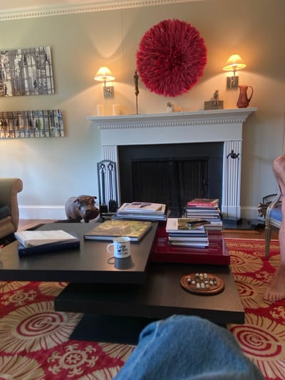 The scene shows a living room interior, likely in a home located in Rye, New York, with a fireplace, a coffee table, and a red rug. The fireplace is white and trimmed with moldings. It has a black metal fire screen and a black hearth. There are a couple of stacks of books on the hearth, and a pair of brass and iron fire tools sit to the left of the fireplace. There are two wall sconces on the wall behind the fireplace, each with a white lampshade. Above the fireplace, a red feather headdress is hanging on the wall. The room also features a black coffee table, a stack of books on it, a white mug with the words "Camp Hun, NY Cats" written on it, a red leather-topped box, and a game board with white tiles. There is a red and gold rug on the floor, and a pair of jeans is in the foreground. A brown hippopotamus statue stands on the floor to the left of the coffee table. There are two framed pictures on the wall to the left of the fireplace. One is a black and white photograph, and the other appears to be a collection of black and white photographs. There are a pair of sconces on the wall in the background. An individual's foot is visible in the right foreground. The overall scene appears relaxed and homey.