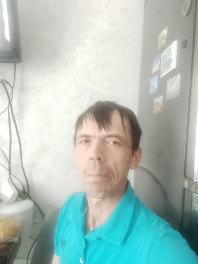 The image depicts a close-up, waist-up view of a man standing indoors, likely in a residential setting in Zyuzyino, Russia. The man has short, dark hair and is wearing a bright blue polo shirt with white buttons. His facial expression appears neutral or slightly downturned. To his left, a television screen is visible, along with coiled black wires and a white electronic device. To his right, a refrigerator is partially visible, adorned with several framed photographs. The background wall has a textured, light-colored finish. The lighting is somewhat dim and diffused, creating a soft, slightly hazy atmosphere.