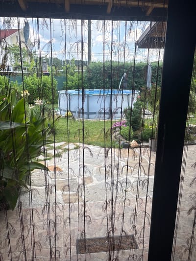 This media file is a photograph taken from indoors, looking out through a beaded curtain at a backyard scene. The backyard features a paved patio with large, light-colored tiles, a patch of green grass, and lush vegetation. In the background, an above-ground swimming pool with blue lining is visible, surrounded by trees and bushes. There is a large house with a red roof on the left side of the frame.
A light-colored cat can be seen on the patio near the grass, and another darker-colored cat is visible in the grass near the pool. There are also some pink and white flowers blooming among the greenery. The sky is partly cloudy, suggesting a pleasant day, likely during daylight hours. The overall scene conveys a peaceful and well-maintained outdoor living space. The beaded curtain in the foreground creates a textured, semi-transparent barrier, adding depth to the image.