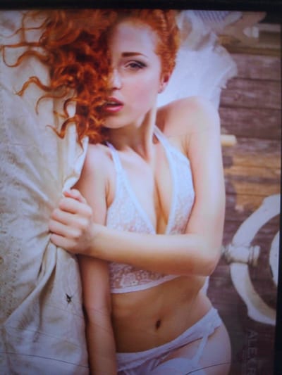 The media file shows a close-up, medium-angle shot of a young woman with long, curly red hair lying on a white linen surface, possibly a bed or a large cushion. She is wearing a delicate white lace bralette and white underwear. Her right arm is bent, and her hand is clutching the fabric of the linen next to her face. Her face is turned to her left, with her eyes looking towards the viewer. Her mouth is slightly open, with a subtle expression of sensuality. The background consists of weathered wooden planks, suggesting an outdoor or rustic setting. There is a circular white object on the right side of the frame, possibly a part of a piece of furniture or a decorative element. The lighting appears soft and diffused, creating a warm and intimate atmosphere. The time of day is not explicitly indicated, but the lighting suggests it could be daytime, possibly morning or afternoon. The overall mood is serene and suggestive. There is partially visible text in the lower right corner of the image, appearing to be "ALEXR", which might be a photographer's or artist's signature.