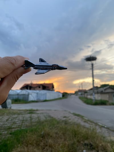 In Miriantsi, Bulgaria, a hand holds a miniature fighter jet in the foreground, with a dusky sky illuminated by a sunset as the backdrop. The jet is black and appears to be made of metal. In the middle ground, a paved road winds through a village. Buildings with red-tiled roofs and some greenhouses with white coverings are visible. A tall utility pole with a large bird's nest at the top stands on the right side of the road. The sky is partly cloudy, with hues of orange, yellow, and gray from the setting sun. The grass in the foreground is dry and patchy.