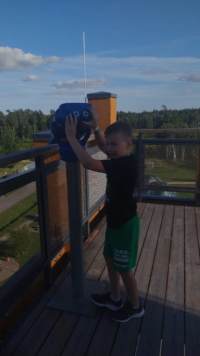 In Tērvete, Latvia, a young boy stands on a wooden platform, looking through a blue public telescope. He is wearing a black t-shirt and green shorts with the words "CREATE, EXPLORE, SURVIVE MINECRAFT" printed on them. The platform has a metal railing with a mesh infill. In the background, a dense forest stretches towards the horizon under a bright blue sky with scattered white clouds. The scene suggests a recreational area or an observation point offering panoramic views of the surrounding landscape.