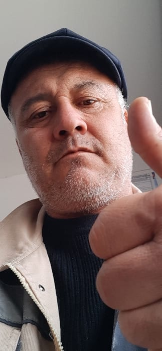The image displays an adult male in a close-up, low-angle indoor shot, located in Qorovulbozor, Uzbekistan. The man, appearing middle-aged to older with grey facial hair, wears a dark blue flat cap, a light beige zippered jacket with a visible snap button near the collar, and a black ribbed garment underneath. He looks directly at the camera with a neutral expression. His right hand is prominently visible in the lower right foreground, forming an upright 'thumbs-up' gesture. The background is a plain light-colored wall, with a blurred, light-colored rectangular object featuring dark markings, consistent with text, partially visible behind the subject's right shoulder.