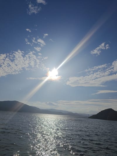 The image depicts a body of water, likely a lake or sea, with a mountainous landscape on the horizon. The sun is high in the sky, partially obscured by clouds, and its rays are creating a strong lens flare effect, with a bright beam extending diagonally across the upper portion of the image. The water is rippled, reflecting the sunlight in a glittering pattern. The sky is a vibrant blue with scattered white clouds. In the distance, dark silhouettes of mountains are visible, and a small navigational marker or lighthouse can be seen on a rocky outcrop. The scene suggests a natural, possibly remote, environment. The location is identified as the city of New Bukhtarma, Kazakhstan, implying this is a view of the Bukhtarma Reservoir. There are no people or animals visible in the image.