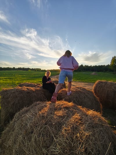 This image depicts a tranquil late afternoon or early evening scene in a rural landscape, likely near Стеклянный, Russia. The warm, golden light of the setting sun bathes the environment, casting long shadows and creating a peaceful atmosphere.

Two individuals are prominently featured, situated on large, round hay bales in the foreground. On the left, a person with light-colored hair, tied up, sits comfortably on a hay bale, seemingly absorbed in her mobile phone. She is dressed in dark attire, her figure silhouetted but her hair catching the sun's glow. To the right, another individual stands atop a different hay bale, viewed from behind. This person, with long, light brown hair, wears a light purple long-sleeved top, light blue denim shorts, and pink sneakers. She faces away from the camera, looking towards the distant horizon and the sun, suggesting a moment of reflection or appreciation of the view.

The hay bales themselves are textured and sun-kissed, forming natural seating and platforms. Beyond the hay bales, a vast, vibrant green field stretches into the midground, eventually meeting a dense treeline that forms the horizon. The sky overhead is a clear, soft blue, adorned with delicate, wispy white clouds, particularly around the sun, which is low in the frame. The overall impression is one of relaxation and connection with nature in a beautiful, open countryside setting. No visible text is present in the media file.