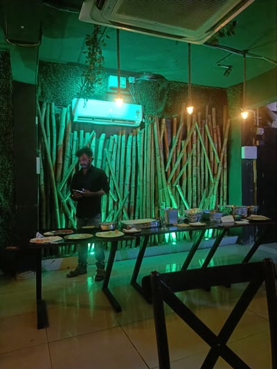A man in a black shirt and jeans, wearing sandals, stands by a table laden with food. He is holding a mobile phone in his hands and appears to be looking at it. The table is set up for a buffet, with various dishes in bowls and on plates. Behind the table is a wall decorated with bamboo stalks and a textured green surface, likely artificial foliage. An air conditioning unit is visible on the wall above the bamboo. Two pendant lights hang from the ceiling, casting a warm glow. The floor is tiled with a reflective surface. The overall lighting in the room is predominantly green, suggesting a stylized ambiance. This scene is likely from a restaurant in Kurnool, India, given the context provided.