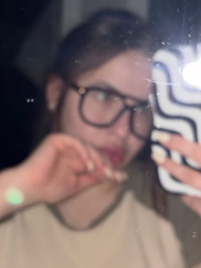 A blurred image shows a person with glasses holding a smartphone with a black and white striped case, seemingly taking a selfie in a dark environment. The person's face is visible, with light skin, dark hair, and a slightly open mouth. A hand is near the person's face. The background is indistinct. The image is marred by blurry spots and glare, hindering precise details. No location information is discernible, so the reference to Shendzhii, Russia, is not supported by the image.