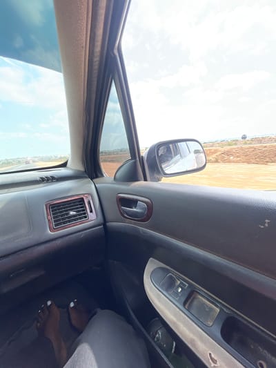 View from inside a vehicle parked in Dokwa, Nigeria. Driver's side door and dashboard visible. The passenger has feet propped on the dashboard. Outside the car is arid land, a few buildings, and a cloudy sky.