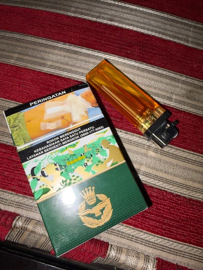 An overhead shot shows a pack of cigarettes and a bright orange lighter on a red and beige striped surface. The cigarette pack is partially open, revealing a graphic image on the front warning about the dangers of smoking. Text on the pack, written in Malay, reads "PERINGATAN" (Warning) and "ROKOK MERENGGUT KEBAHAGIAAN SAYA SATU PERSATU. LAYANAN BERHENTI MEROKOK (0800-177-6565)" which translates to "Smoking steals my happiness one by one. Quit Smoking Service (0800-177-6565)". The bottom part of the pack displays a green design with gold embellishments, including a crown and laurel wreath. The lighter is positioned to the right of the cigarette pack, with its transparent orange casing revealing the internal mechanism. The striped surface appears to be fabric, possibly a rug or upholstery. The lighting suggests an indoor environment, with the angle of the light creating some shadows. The specific location in the city of Tupah, Indonesia, is not discernible from the visual information provided.