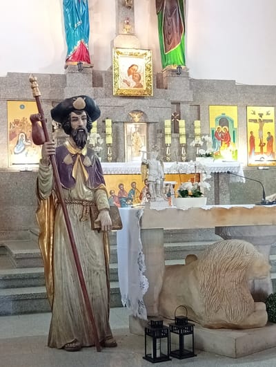 The image depicts the interior of a church in Sobótka, Poland, specifically within the Holy Trinity Parish Church. In the foreground, a life-sized statue of Saint James the Greater stands prominently. The saint is depicted as a pilgrim, wearing a wide-brimmed hat adorned with a scallop shell, a purple cloak over a golden robe, and carrying a staff and a book. Behind the statue, the church altar is visible, decorated with religious icons and candles. To the right of the altar, a stone sculpture of a lion rests on a pedestal, and in front of it, two black metal lanterns with candles are placed on the floor. The church's architecture features stone elements and a light-colored wall in the background.