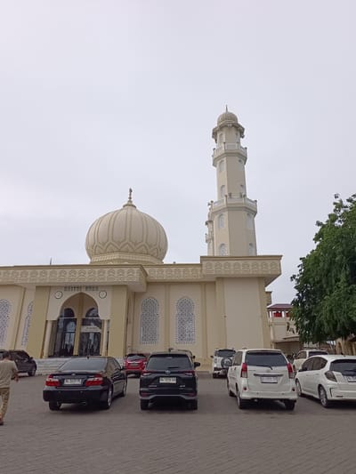 In the city of Mee Tanjong Usi, Indonesia, a mosque named "BAITUL HUDA" is depicted. The mosque features a prominent cream-colored dome and a tall, slender minaret. The building's facade is adorned with decorative patterns and arched windows. In front of the mosque, a paved courtyard is filled with several parked cars, including a black sedan, a black SUV, a white minivan, and a white hatchback. A lone person, dressed in light-colored pants and a tan shirt, is walking away from the viewer in the foreground. The sky is overcast, suggesting a cloudy day. Lush green trees are visible to the right of the mosque.