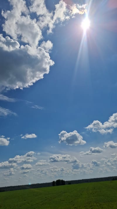 The image shows a vast, bright blue sky filled with scattered white cumulus clouds. The sun is prominently visible in the upper right corner, casting bright rays and lens flare. Below the sky, a rolling green field occupies the lower portion of the frame, with a dark line of trees forming a distant horizon. There are no people or animals visible in the image. The scene appears to be a rural landscape, likely in a location such as Stantsionno-Oyashinskii, Russia, characterized by open fields and clear skies.