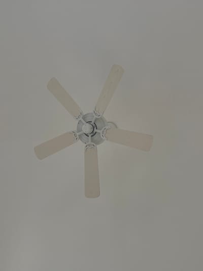A ceiling fan dominates the scene, centered against a plain, grayish-white background. The fan is white, featuring five blades evenly spaced around a central hub. The blades exhibit a subtle wood-grain texture. The lighting is soft and diffused, contributing to the overall muted tone of the image.