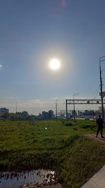The image depicts a sunny day in Obukhovo, Russia. The sun is high in a clear, light blue sky. A grassy area runs along the right side of the frame, adjacent to what appears to be a road or highway, with occasional standing water reflecting the light near the edge. Utility poles and some kind of steel structure run along the right side of the road, adjacent to the field. A person is walking on the sidewalk along the right edge of the image. Trees and distant buildings are visible on the horizon. Several cars can be seen on the road.