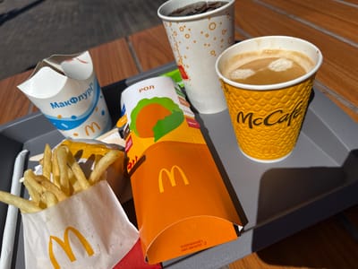 A McDonald's tray sits on a wooden table outdoors. On the tray are a white paper cup with a design and the "McDonald's" logo, a white paper bag with french fries and the "McDonald's" logo, an orange box with the "McDonald's" logo and a graphic of food, a white cup with orange circles and the "McDonald's" logo, and a yellow cup with a textured pattern and the "McCafé" logo. The McCafé cup contains coffee with foam. The white cup contains a dark beverage with ice. The overall scene suggests a meal purchased from McDonald's. The image was taken in Hadzynka, Ukraine.