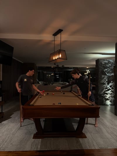 Two young men are playing billiards in an indoor room. One man in a dark long-sleeved shirt is leaning over the pool table pointing towards a ball. The other man, wearing a grey shirt and dark pants, is holding a pool cue as if preparing to take a shot. The pool table is wooden and has several billiard balls scattered on its surface. Above them is an industrial-style light fixture with visible bulbs. The room has a stone feature on one wall. One person is partially sitting on the pool table. The exact location in Santa Ana Yancuitlalpan (Chilhuacán), Mexico, is indeterminate.