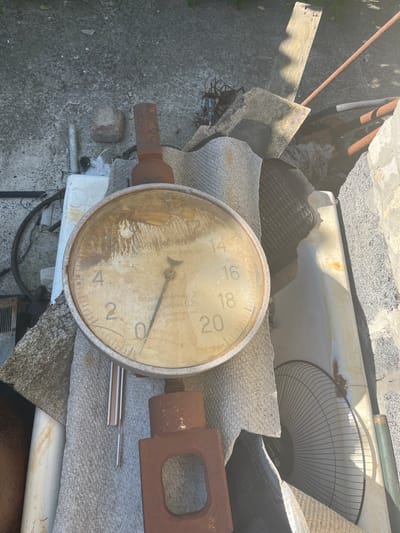 This is a close-up, outdoor, overhead shot of a collection of objects, possibly in a yard or storage area, in Bilenke, Ukraine. The main subject is a large, round gauge with a faded, yellow face and a metal rim. The gauge has markings and numbers, including "14", "16", "18", and "20", and a needle pointing between "2" and "4". There is visible text on the gauge face, including what appears to be Cyrillic script.
Surrounding the gauge are various other items. To the left of the gauge, a rusty metal rod extends diagonally across the frame. Several pieces of weathered wood and gray, textured material are also present. To the right of the gauge, a rusty metal bracket with a rectangular opening is visible. Beneath the gauge, there's a white plastic object and some black cabling. In the background, elements like concrete, rusty pipes, and a dark, patterned material (possibly a grate or mat) can be discerned. The scene appears to be lit by natural daylight, suggesting it is daytime, and the overall impression is one of disuse and accumulation of various discarded items. There are no people visible in the image.