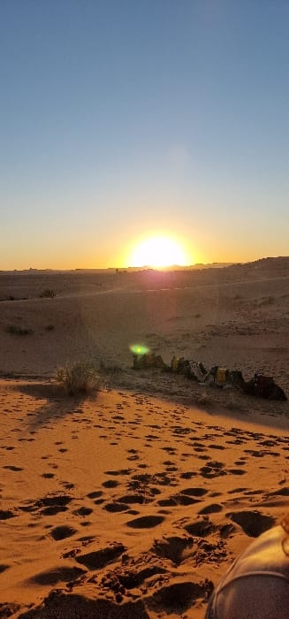 A vertical photograph captures a desert landscape at either sunrise or sunset in Et-taous, Morocco. The large, bright disc of the sun is partially visible on the central horizon, casting a luminous glow and a prominent lens flare. The sky transitions from a deep blue at the top to a bright orange-yellow at the horizon. The midground features undulating sand dunes with sparse, low-lying desert vegetation. Approximately six dromedary camels, equipped with yellow and brown saddles, are observed lying prone on the sand, positioned to the right of the center. Distant structures or hills are faintly silhouetted against the bright horizon. The foreground consists of reddish-orange sand, heavily marked with numerous human footprints. A partial view of a human leg or foot is visible in the lower right corner, indicating a person is seated or reclining on the sand while capturing the image.
