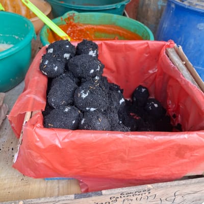 In Mesjid Tuha, Indonesia, a close-up, eye-level shot captures a red plastic-lined cardboard box filled with salted duck eggs. The eggs are coated in a dark, earthy mixture, with some showing white patches of salt. The background is out of focus, but appears to be a market stall with various containers, including green buckets containing reddish-orange food items and a large blue barrel. A green plastic utensil is visible in the upper left quadrant. The overall impression is one of a food preparation or display setting within a market environment.